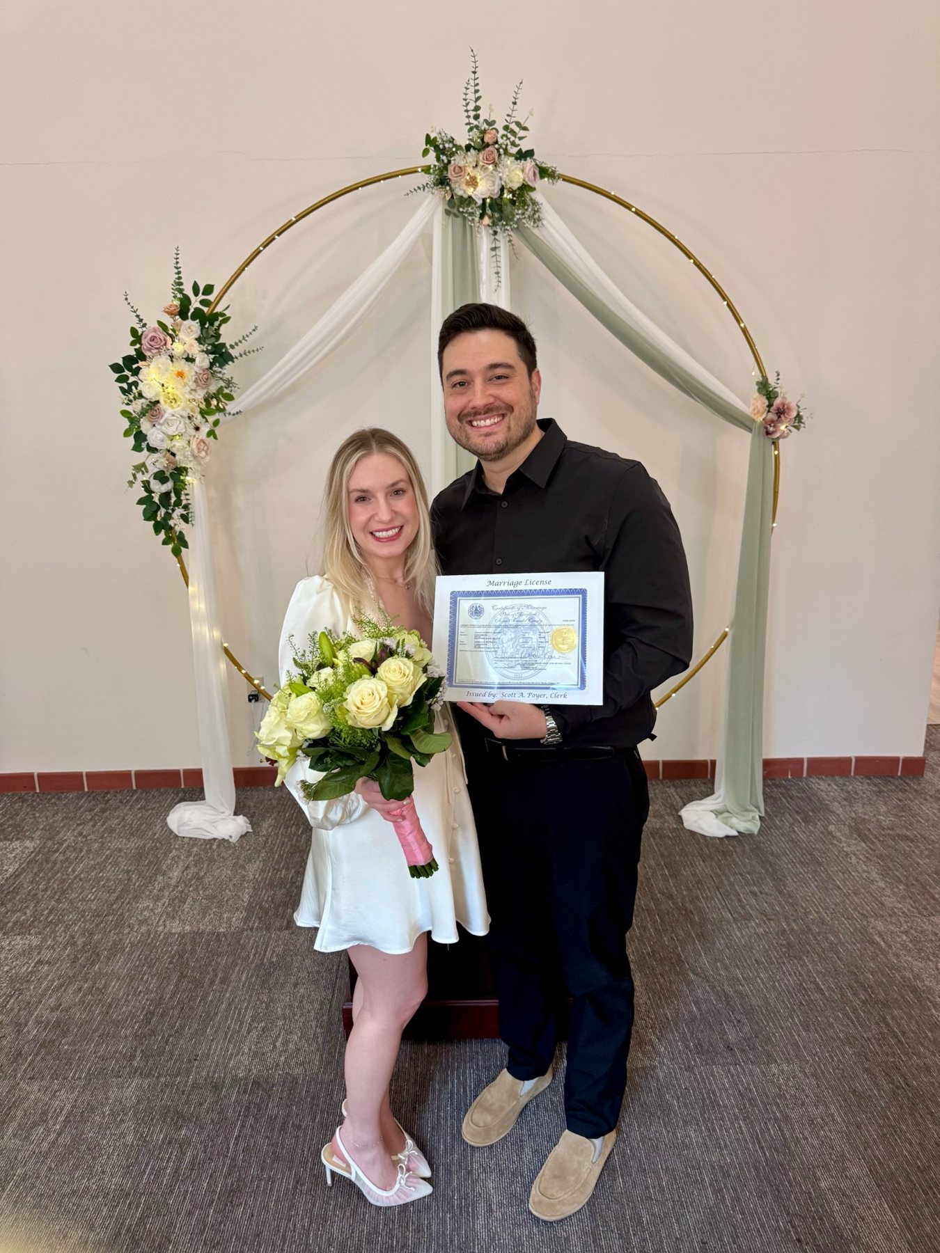 Andrea and Vince just married, holding their marriage license under the floral arch